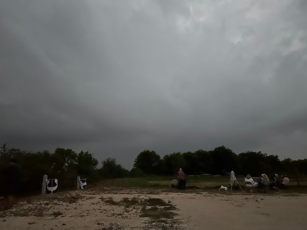 Dark gray sky, silhouette of trees, and a few scattered people on sandy soil.